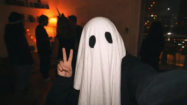 Person taking a flash selfie at a dark Halloween party wearing a white ghost sheet with two black eyeholes
