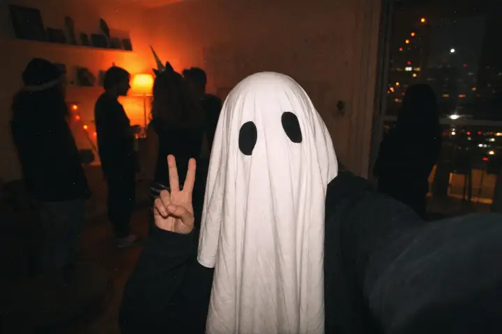 Person taking a flash selfie at a dark Halloween party wearing a white ghost sheet with two black eyeholes