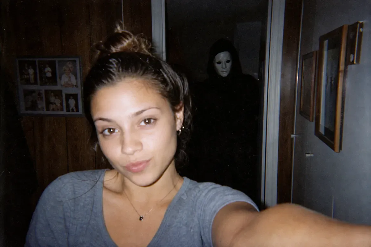 A selfie in a dim 1990s hallway with a faint pale-masked figure standing in the doorway behind the subject