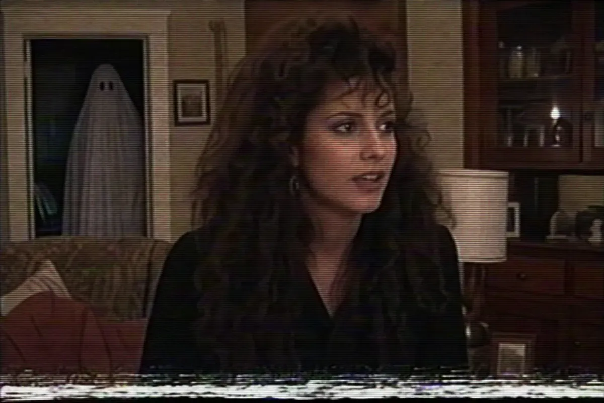 VHS frame-grab style horror still with scan lines and a bedsheet ghost in the background doorway